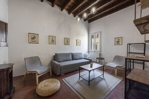 A living room with a grey sofa, two armchairs, a coffee table, and a woven ottoman. There are framed pictures on the wall, a window with shutters, and a wooden ceiling. - Ruote Nest by MMega Homes and Villas  (Florence)