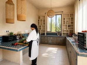 A kitchen with a person chopping vegetables, a microwave, and a large window. - Palais Samalya (Bouzama)