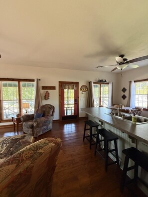 A spacious living area with a wooden floor, a large window, a ceiling fan, and a kitchen area with a bar counter and stools. - The Cabin on Monkey Island (Afton)