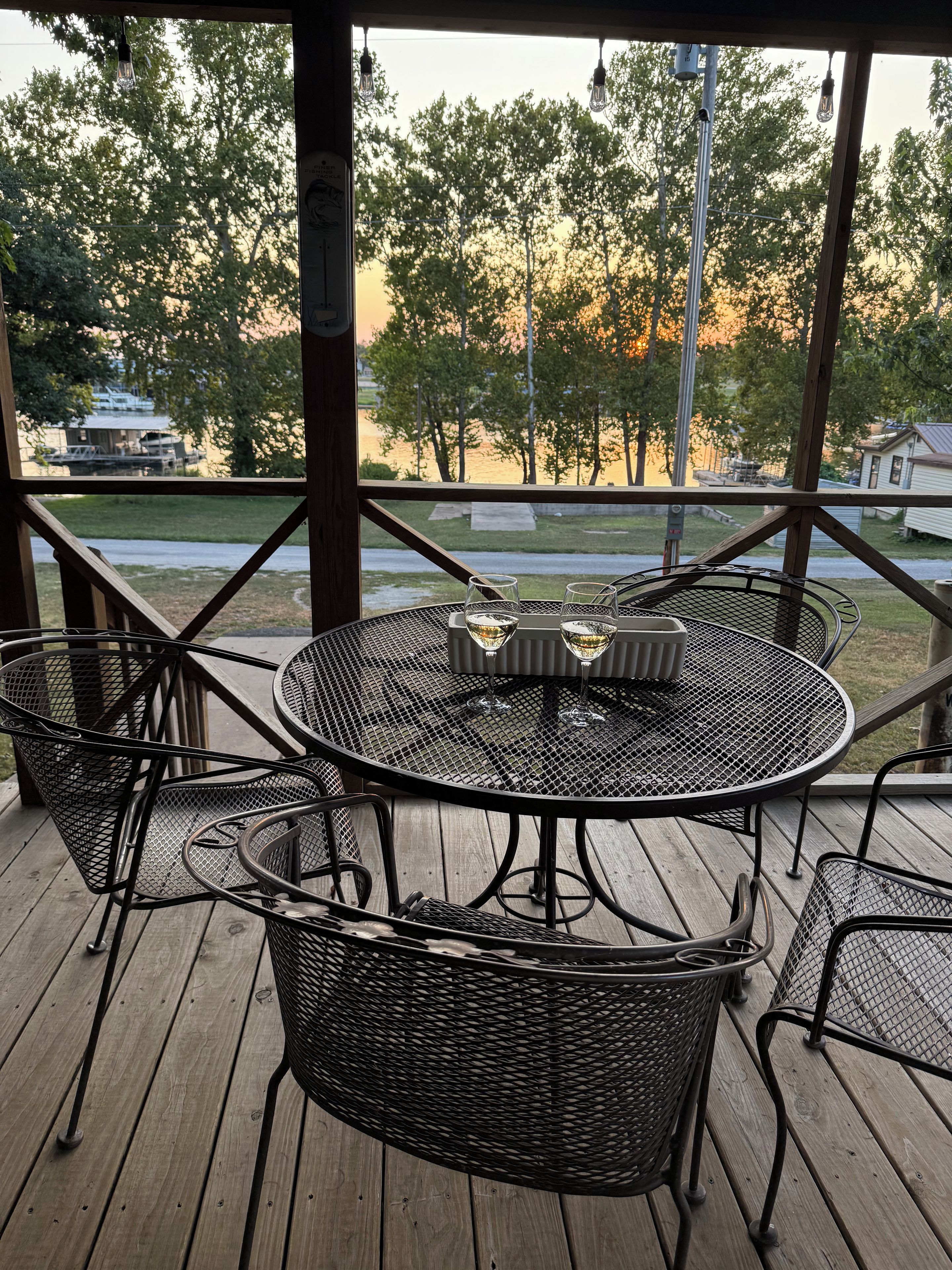 A covered outdoor seating area with a round metal table and chairs, overlooking a body of water and trees.