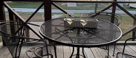 A covered outdoor seating area with a round metal table and chairs, overlooking a body of water and trees.