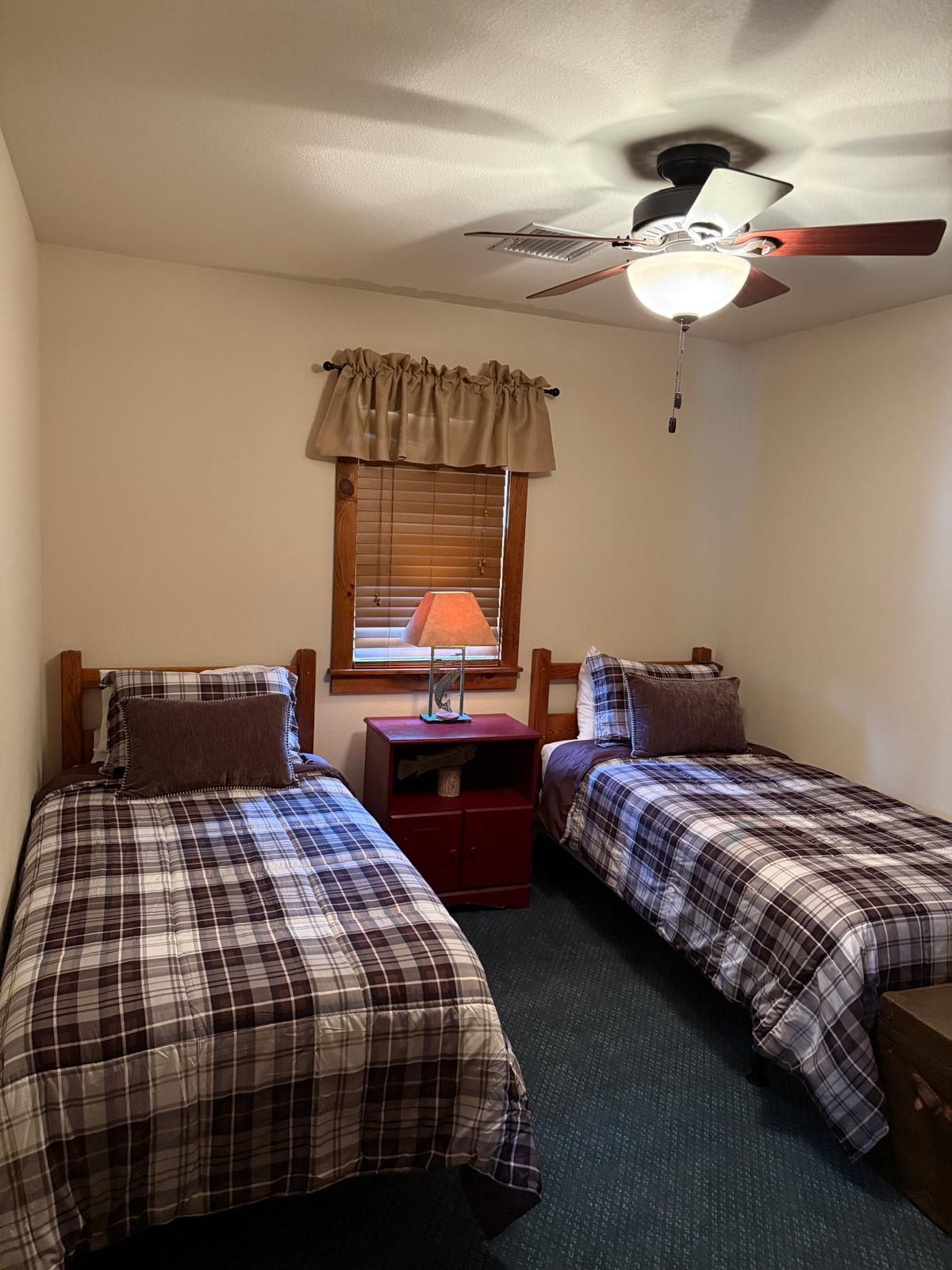 A bedroom with two beds, a ceiling fan, a window with blinds, and a bedside table with a lamp.