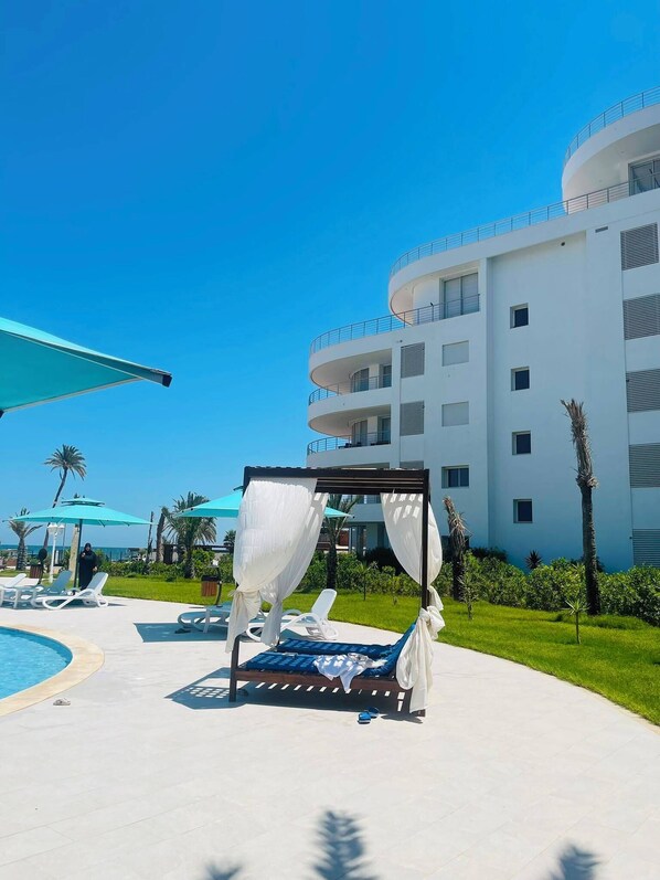 A resort pool area with a cabana, lounge chairs, and a large building in the background.