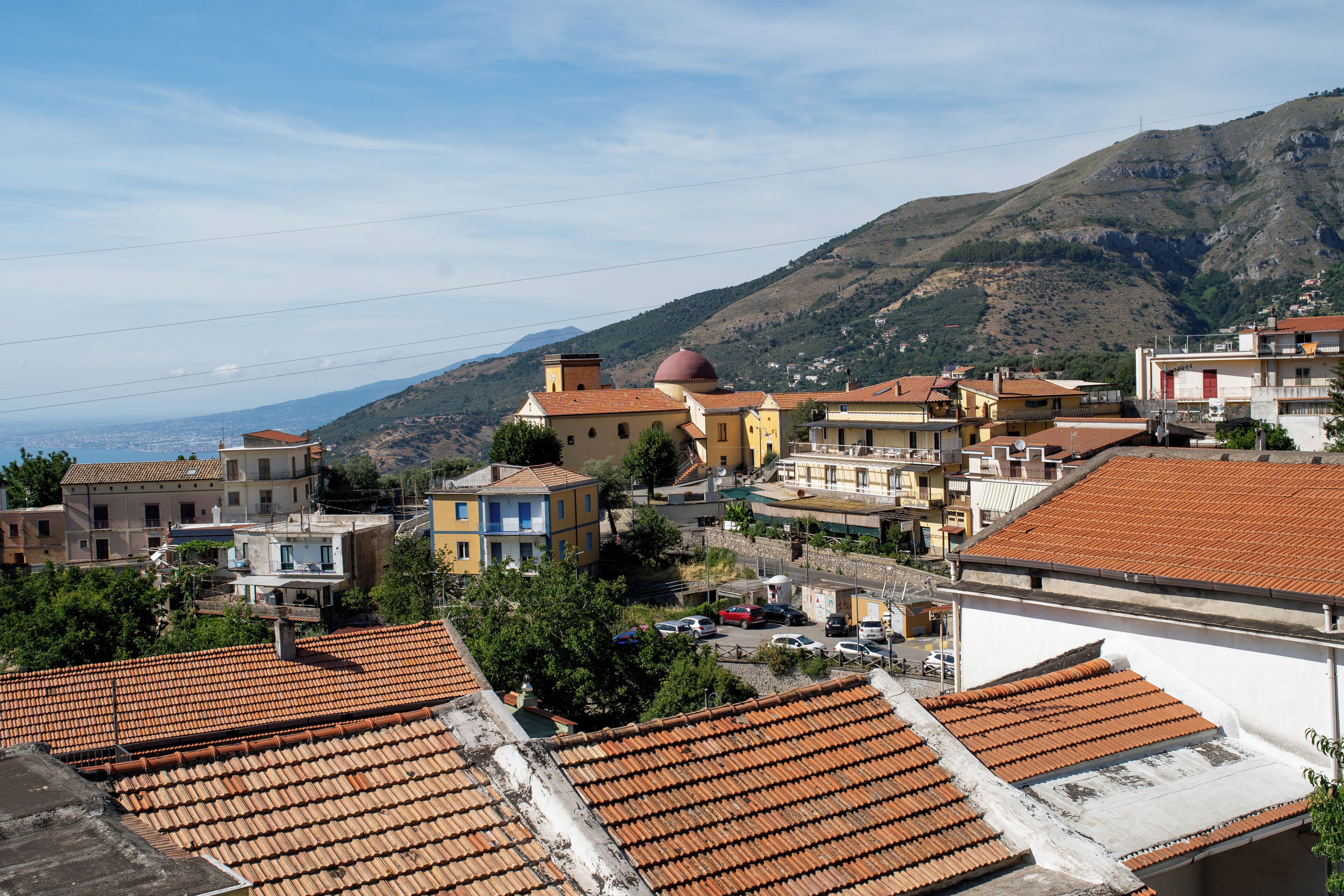 A coastal town with terracotta-roofed buildings, a prominent church with a dome, and a view of the sea and mountains in the background.