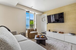 A modern living room with a sofa, armchair, and coffee table, a flat-screen TV mounted on the wall, and a view of the outdoors through a sliding door. - Lavie Maison | Le Marais & Rivoli (Paris)