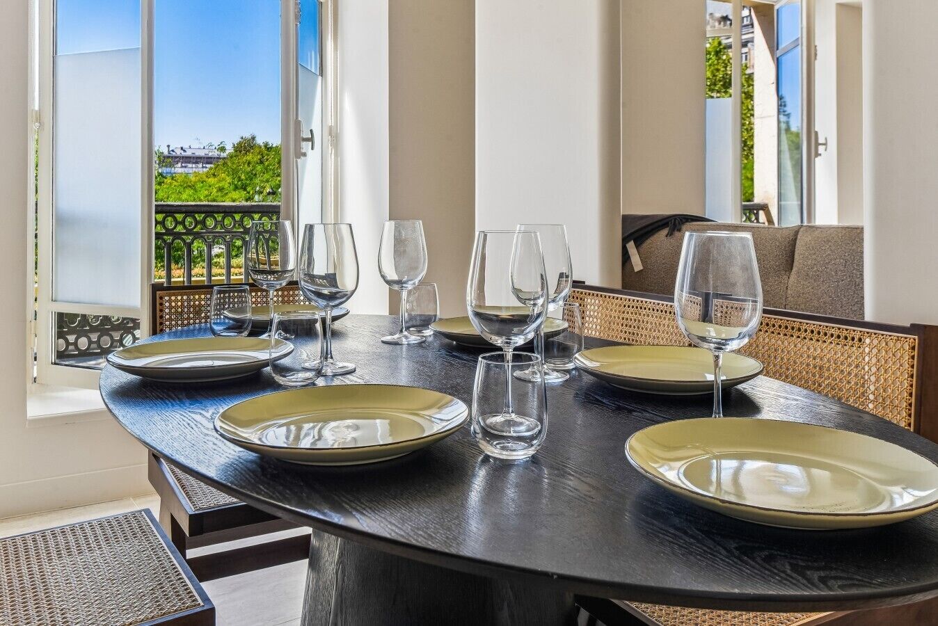 A dining table set with plates and wine glasses, with a view of greenery and buildings outside.