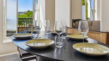 A dining table set with plates and wine glasses, with a view of greenery and buildings outside.