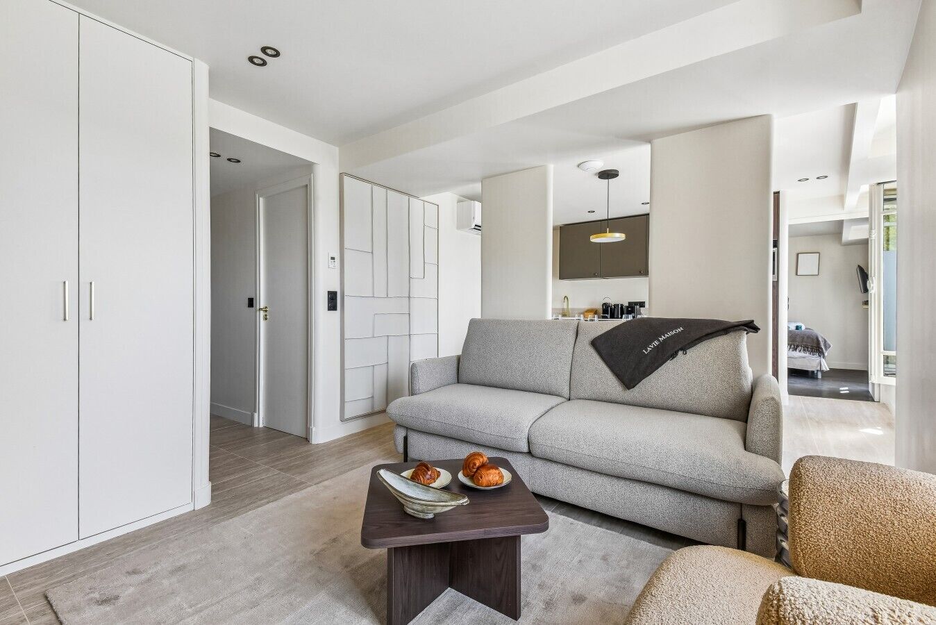 A modern living room with a grey sofa, a coffee table with pastries, and a kitchen area in the background.