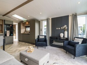 A modern living room with a sofa, ottoman, and a fireplace. There is a kitchen area with appliances and a dining table. - Lakewood Plot 8 (Clacton-on-Sea)