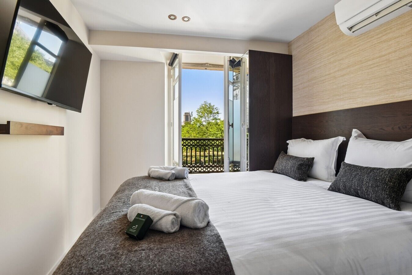 A modern hotel room with a large bed, a flat-screen TV mounted on the wall, and a balcony with a view of the city.