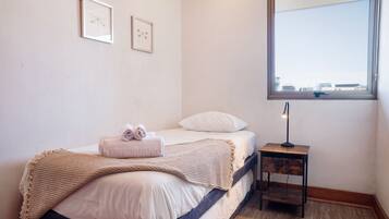Un dormitorio con cama, una mesita de noche con lámpara, una ventana con vistas, y dos cuadros en la pared.