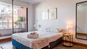 Ein modernes Hotelzimmer mit einem großen Bett, einem Balkon mit Blick auf Gebäude, einem kleinen Tisch mit einer Lampe und zwei gerahmten Pflanzenbildern an der Wand.