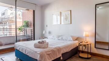 Habitación de hotel moderna con una cama grande, un balcón con vistas a edificios, una mesita con una lámpara y dos láminas botánicas enmarcadas en la pared.