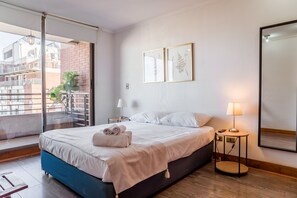 A modern hotel room with a large bed, a balcony with a view of buildings, a small table with a lamp, and two framed botanical prints on the wall. - Cozy Getaway Oasis 2BR in Las Condes (Las Condes)