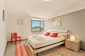 A bedroom with a bed, a nightstand, a chair, a window with a view, and a painting on the wall. - [SanVenerio] Sea view 5 TERRE (La Spezia)