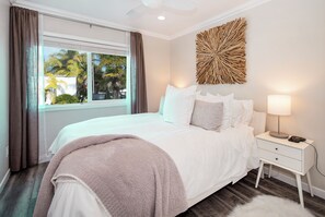 A bedroom with a bed, a nightstand, a lamp, a window with curtains, and a wall decoration.