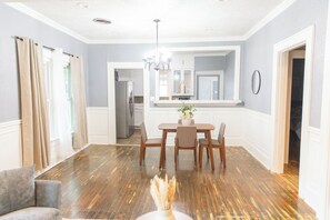 Uma sala de jantar com mesa e cadeiras de madeira, um lustre e vista para a cozinha.