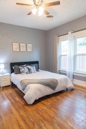 Quarto com cama, mesa de cabeceira, ventoinha no teto e janela com cortinas.