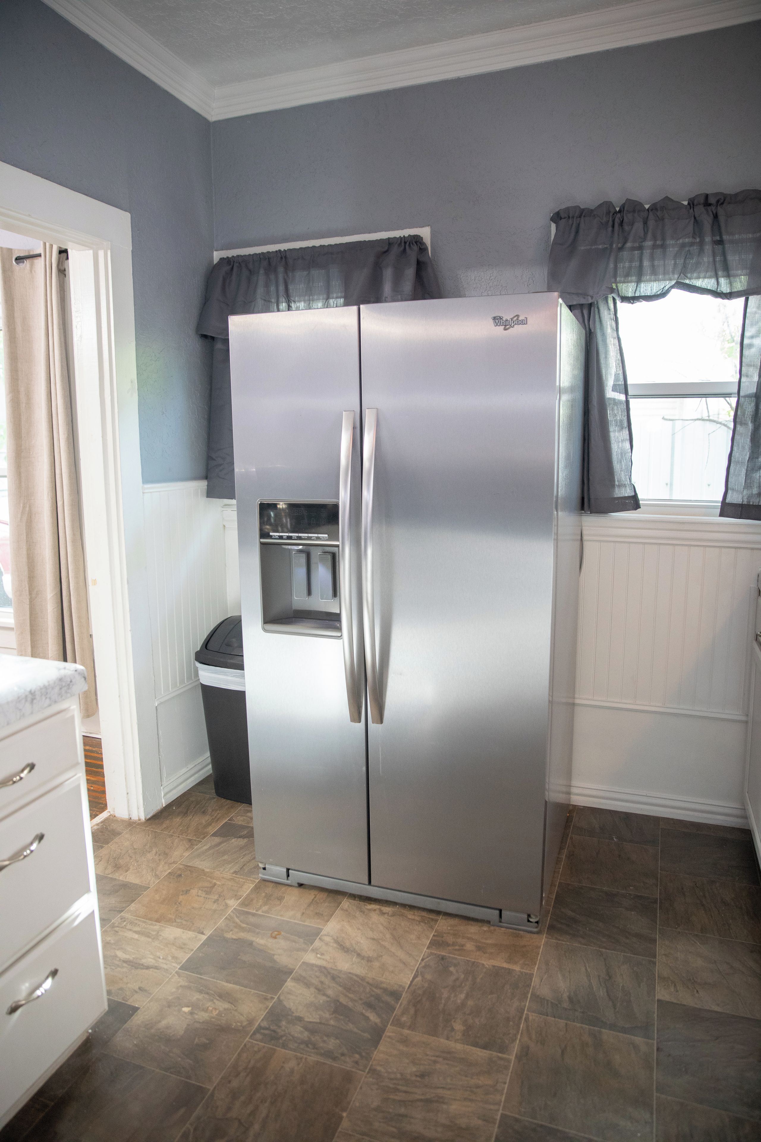 A stainless steel Whirlpool refrigerator with a freezer compartment, located in a kitchen with a tiled floor and a window with curtains.