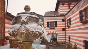 A stone urn with a lion's head on a pedestal, surrounded by potted plants, in a courtyard with striped buildings.