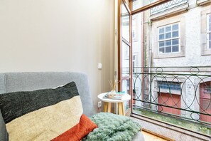 Ein Zimmer mit einem Bett, einem kleinen Tisch mit einer Zeitschrift und Blick auf ein Gebäude durch ein Fenster.