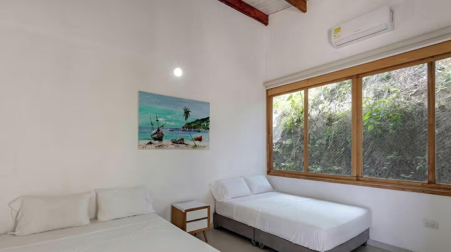 A room with a bed, a small bedside table, a window with a view of greenery, and a painting of a beach scene.