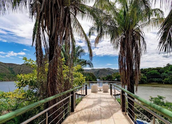 A bridge with a railing leading to a seating area overlooking a body of water and hills.