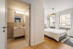 Quarto moderno com cama grande, mesa de cabeceira, janela com vista para edifícios e banheiro com pia e espelho.