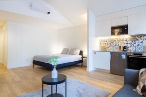 A compact room with a bed, a small table with a vase, a kitchenette with a microwave, and a grey sofa. - Historic Porto Studio Steps from Clérigos Tower (Porto)