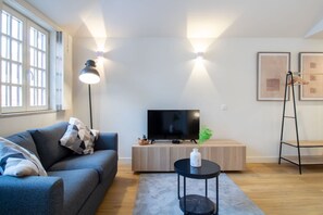 A modern living room with a sofa, a TV on a wooden stand, a small table with a vase, and a floor lamp. - Historic Porto Studio Steps from Clérigos Tower (Porto)