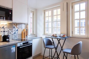 A compact kitchen with a microwave, oven, and a small refrigerator. There are two bar stools and a table with a cup and a plate. - Caldeireiros 95 G by Astoundinn (Porto)