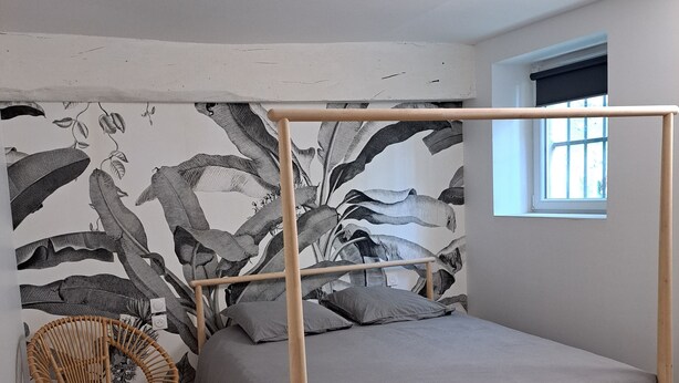 Een enkel bed met houten frame en grijze beddengoed, een rieten stoel en een wand met een groot botanisch kunstwerk.