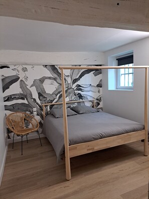 Uma cama de solteiro com estrutura de madeira e roupa de cama cinza, uma cadeira de vime e uma parede com uma impressão botânica grande.
