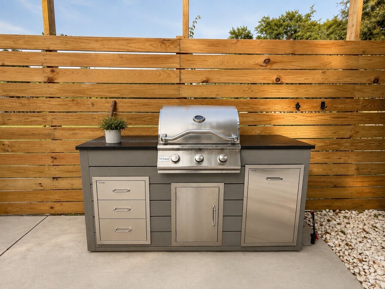Een buitenkeuken met een ingebouwde grill, opbergkasten en een kleine plant erboven.