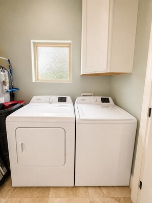 A laundry room with two white front-loading washers and dryers, a window above them, and a cabinet above the dryers. - Fully Renovated Beach Retreat in Sunnyside (Panama City Beach)