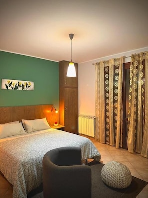 A bedroom with a bed, a chair, a bedside table with a lamp, a radiator, and patterned curtains.