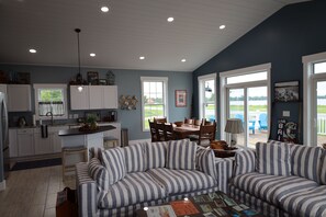 A modern living room with a striped sofa set, a dining area with wooden chairs, and a kitchen with white cabinets. - NEW—The Sandee Gables Cottage at Indian Lake (Lakeview)