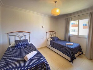 A room with two beds, each with a metal headboard and a blue star-patterned bedspread. There is a window with curtains, and a single light fixture on the ceiling. - Los Corales 1 - Four Bed Townhouse (Arona)