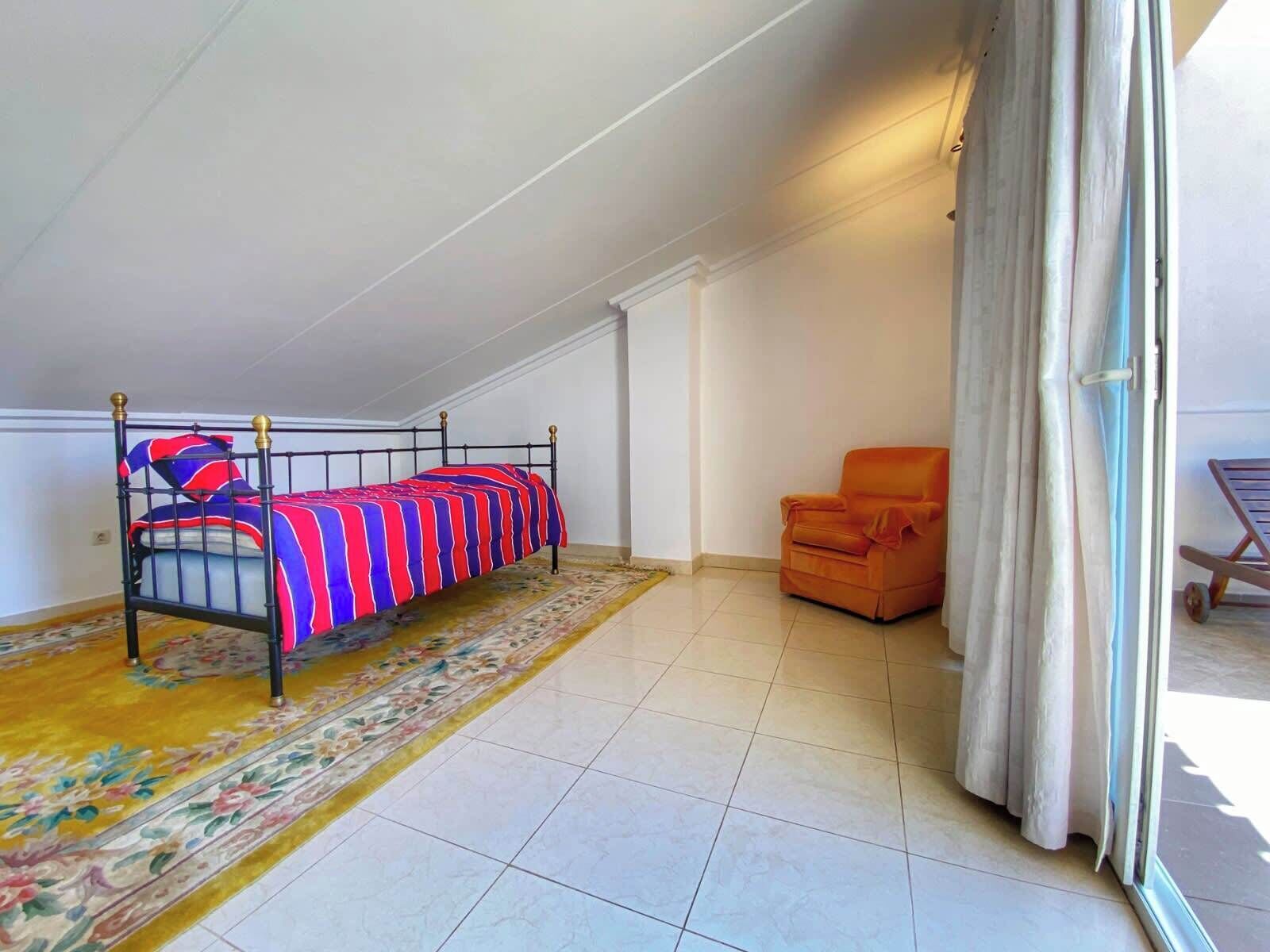 A room with a bed, an orange chair, a window with curtains, and a patterned rug.
