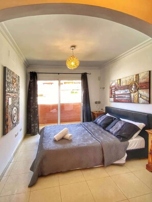 Ein Schlafzimmer mit einem Bett, einer Hängelampe, einem Fenster mit Vorhängen und einem Wandbild.