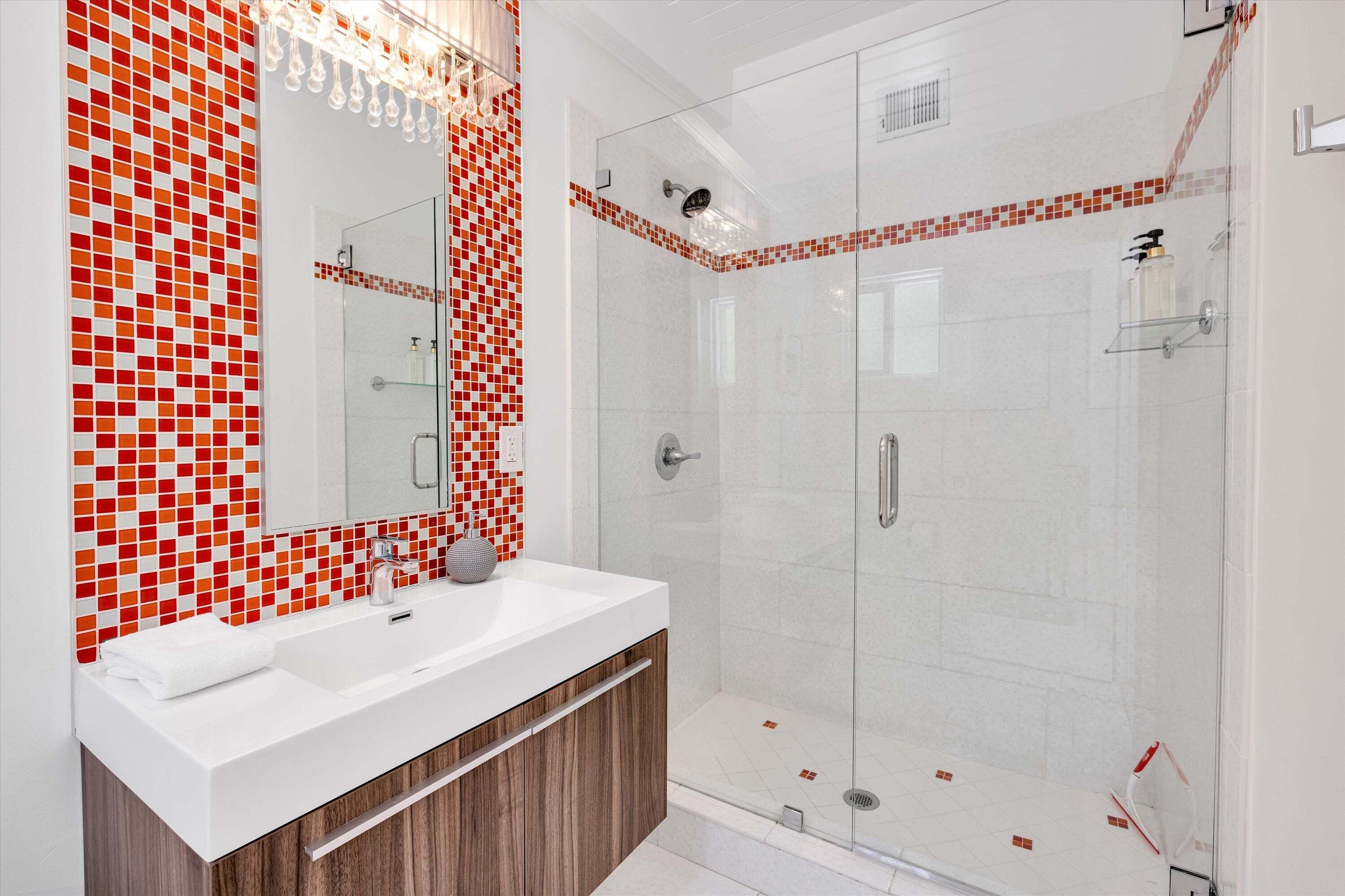Une salle de bain moderne avec une douche à parois de verre, un lavabo blanc et un miroir agrémenté d’un pare-splash en carreaux rouges.