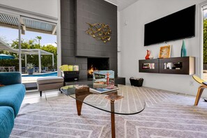 Ein modernes Wohnzimmer mit Kamin, Fernseher an der Wand und Pool, der durch die Glastüren sichtbar ist.