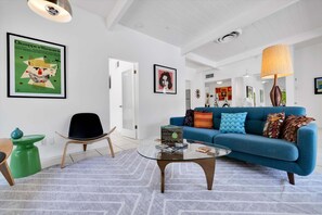 Ein modernes Wohnzimmer mit einem blauen Sofa, einem Glastisch und einem grünen Sessel. An den Wänden befinden sich gerahmte Kunstwerke und auf dem Sofa steht eine Lampe.