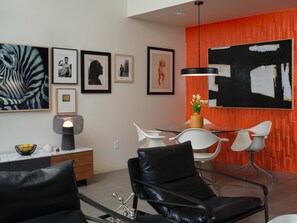 A modern living room with a dining area, featuring a glass table, white chairs, a black leather sofa, and artwork on the walls. - Discover Sophisticated Desert Living at Sophia at Cerritos (Palm Springs)