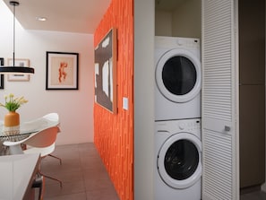 A modern laundry room with two front-loading washing machines, a dining area with a white table and chairs, and a wall-mounted artwork. - Discover Sophisticated Desert Living at Sophia at Cerritos (Palm Springs)