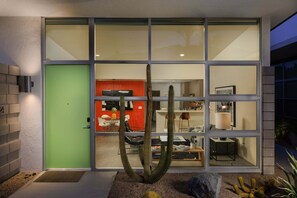 Ein modernes Haus mit einem großen Glasfenster, einem Kaktus im Vorgarten und einem roten Innenraum, der durch das Fenster sichtbar ist.