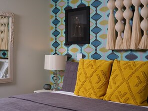 A bed with a gray blanket, a lamp, and two pillows, one purple and one yellow, with a patterned wall behind. - Discover Sophisticated Desert Living at Sophia at Cerritos (Palm Springs)