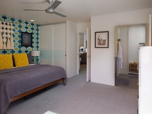 Une chambre à coucher avec un lit, un ventilateur de plafond, une armoire et une salle de bain visible à travers une porte ouverte.