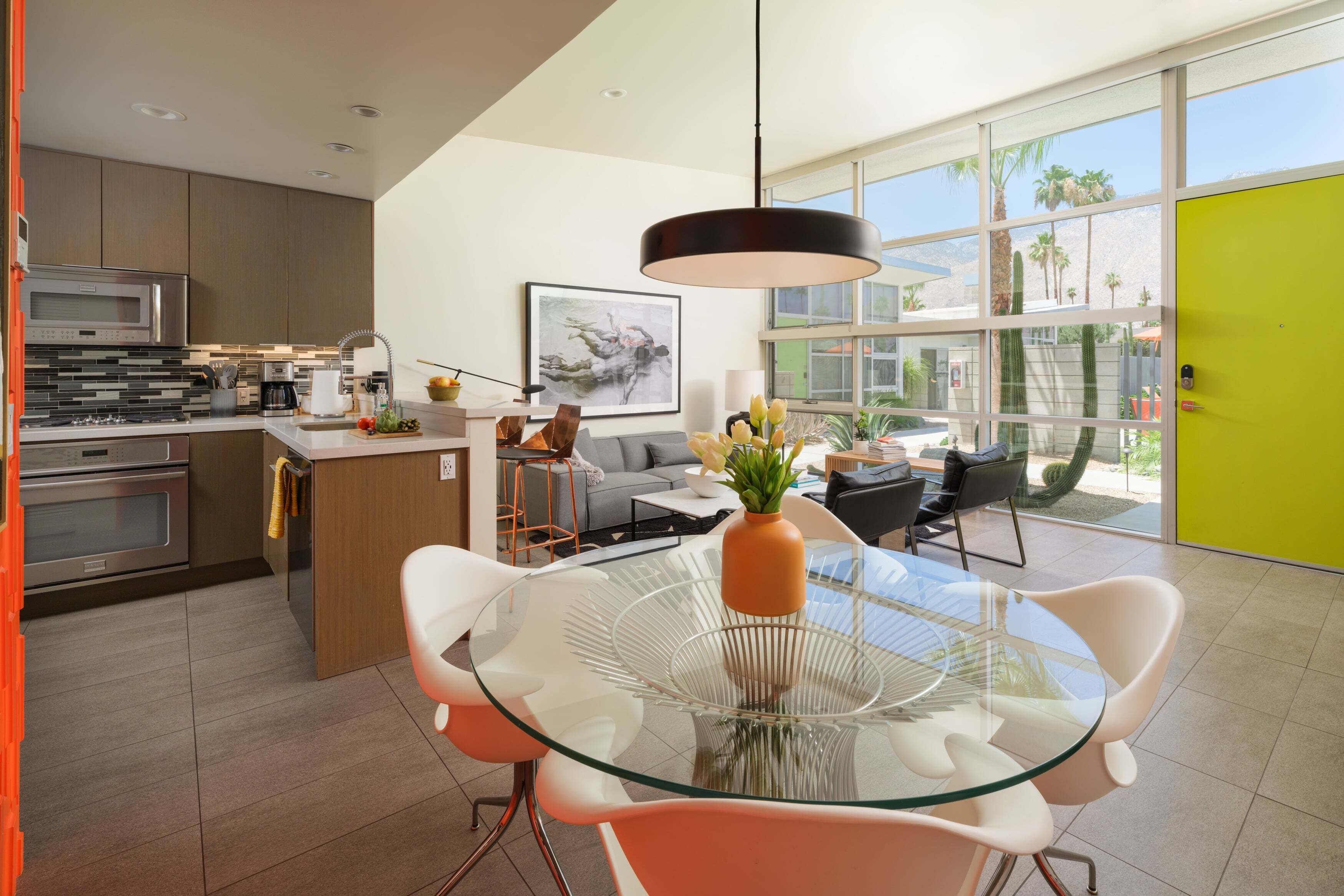 A modern kitchen with stainless steel appliances, a dining area with a glass table and orange chairs, and a view of palm trees through large windows.