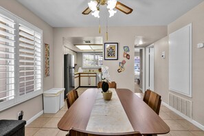 A dining area with a long table, chairs, a ceiling fan, and a kitchen in the background. - Welcome to Mesquite Breeze, Your Relaxing Palm Springs Getaway (Palm Springs)